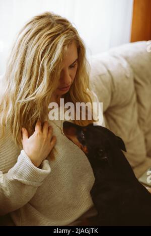 Ritratto di giovane donna con capelli lunghi e ondulati con maglione beige, seduta sul divano, guardando il cane Zwergpinscher. Foto Stock