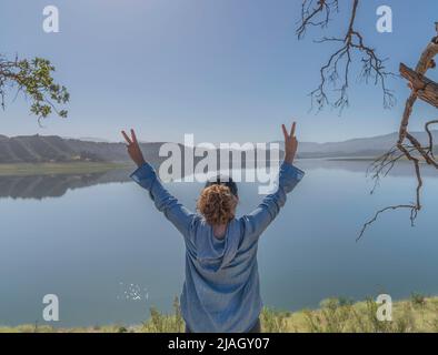 Una donna atletica alza le mani in testa e dà un doppio segno di pace al lago Cachuma, nella contea di Santa Barbara, California. Foto Stock