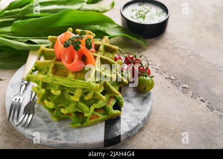 Waffle belgi verdi. Spinaci o aglio selvatico o pesto cialde con salmone rosso e salsa di panna su sfondo grigio tavolo di cemento. Deliziosa colazione, Foto Stock