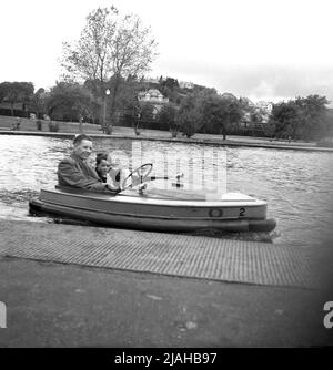 1950s, storica Gran Bretagna del dopoguerra, su un lago in barca in un parco, un padre con il suo giovane figlio fuori insieme, in una piccola barca di legno elettrico da piacere dell'epoca. All'inizio del 20th secolo, i laghi in barca cominciarono ad essere costruiti come forma di svago per le persone che vivono nelle città. All'inizio del 1930s, alcune barche a remi venivano sostituite da imbarcazioni con nuove tecnologie, piccole imbarcazioni a motore elettrico, con una velocità di 4mph, la prima delle quali era la 'Glida', presso il Parco Battersea nel 1929. Sono state introdotte diverse versioni di questo piccolo motoscafo elettrico che sono rimaste in uso fino al 1960s. Foto Stock