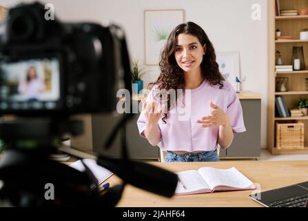 Giovane donna caucasica seduta alla reception di fronte alla macchina fotografica parlando su argomento durante la registrazione di video per il blog Foto Stock