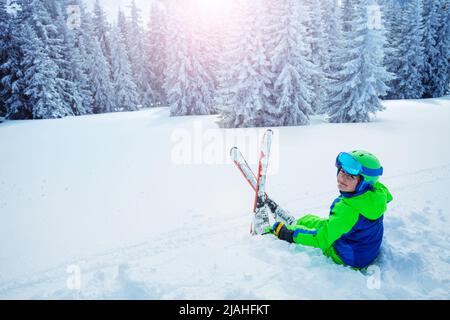 Il bambino si siede nella neve sorridendo sopra la foresta di abeti dopo la nevicata Foto Stock