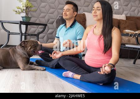 Una giovane coppia di Latino la cui moglie è incinta pratica la meditazione nel loro soggiorno con il loro bulldog accanto a loro sul pavimento Foto Stock