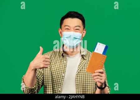 Ritratto di turista maschile asiatico maturo in maschera di tenuta passaporto con biglietti, mostrando pollice su, sfondo verde Foto Stock