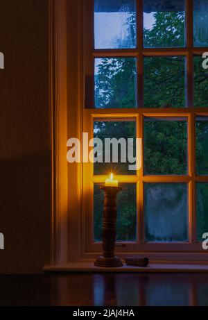 Una candela illuminata in un portacandele di legno vicino ad una finestra durante la luce bassa Foto Stock