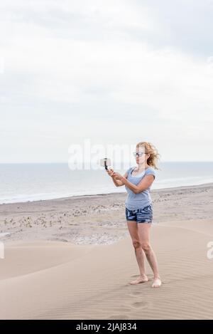 Donna matura con capelli biondi indossando occhiali da sole scattando una foto con smartphone sulla parte superiore di una duna. Spiaggia e mare dietro. Foto Stock