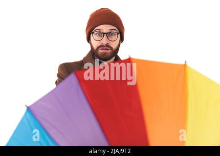 Ritratto da studio di un uomo hipster caucasico in abiti autunnali tra cui una beania e un cappotto che gira il suo ombrello arcobaleno con un'espressione del viso sorpresa. Foto di alta qualità Foto Stock