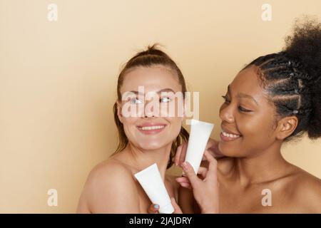 Donne africane ed europee con moisturizer. Felice diverse giovani donne godendo la morbidezza di pelle pulita idratata, spa industria su sfondo beige Foto Stock