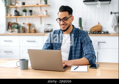 Bell'imprenditore maschile indiano o arabo positivo, freelancer, lavorando da remoto mentre si siede a casa in cucina, utilizzando il computer portatile, digitando sulla tastiera, sviluppare progetto creativo di successo, sorridente Foto Stock