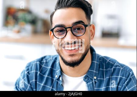 Primo piano ritratto di un bel carismatico arabo o indiano con occhiali, freelance o studente, in abiti casual, con denti bianchi perfetti, guardando la macchina fotografica con un sorriso allegro e noioso Foto Stock