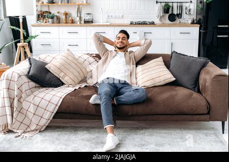 Calmo ragazzo indiano o arabo felice con gli occhi chiusi, mettere le mani dietro la testa, seduto in un moderno soggiorno su un comodo divano, godere di calma e da solo a casa nel fine settimana, sognando, sorridendo Foto Stock