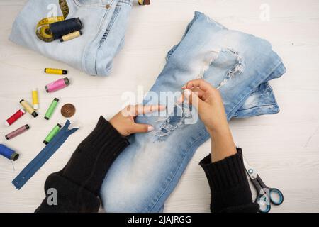 dettaglio delle mani cucendo un paio di pantaloni con un ago e filo, tavolo da lavoro del comò, utensili da lavoro in studio Foto Stock