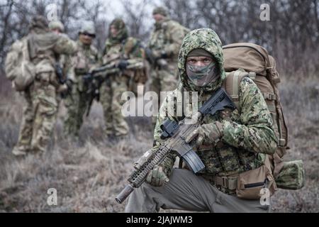 Soldato di fanteria che indossa una giacca camo con cappuccio e uno zaino, inginocchiandosi con l'arma da fuoco nelle mani. Foto Stock
