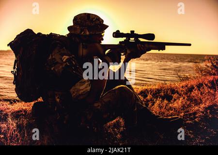 Silhouette di un fucile da cecchino mentre si siede sulla riva dell'oceano durante il tramonto. Foto Stock