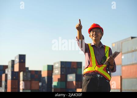 Un ingegnere anziano anziano di lavoratori asiatici che indossa un gilet di sicurezza e un casco in piedi e che tiene un tablet digitale nel cantiere di spedizione dei contenitori di carico. Foto Stock
