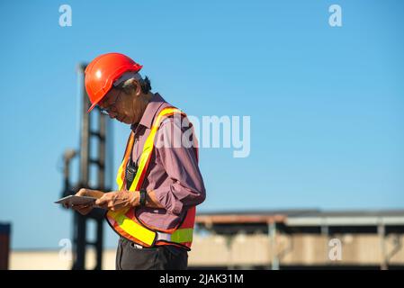 Un ingegnere anziano anziano di lavoratori asiatici che indossa un gilet di sicurezza e un casco in piedi e che tiene un tablet digitale nel cantiere di spedizione dei contenitori di carico. Foto Stock