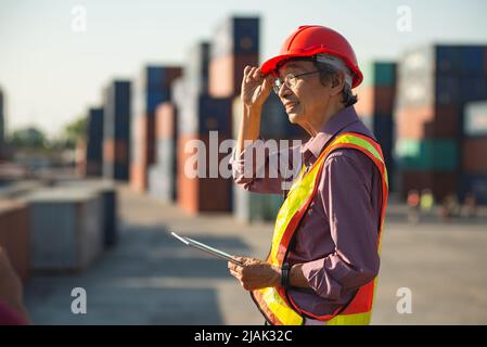 Un ingegnere anziano anziano di lavoratori asiatici che indossa un gilet di sicurezza e un casco in piedi e che tiene un tablet digitale nel cantiere di spedizione dei contenitori di carico. Foto Stock