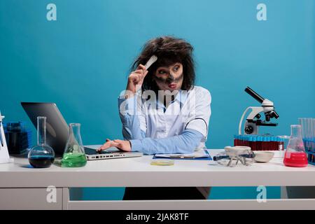 Stolto goofy biochimica esperto graffiare testa cercando di capire perché l'esperimento chimico ha portato all'esplosione. Scienziato lunatico confuso con look selvaggio seduto alla scrivania in laboratorio. Foto Stock