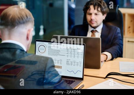 Persone aziendali serie che esaminano il marketing finanziario su notebook con grafici sul monitor mentre lavorano al tavolo in ufficio Foto Stock