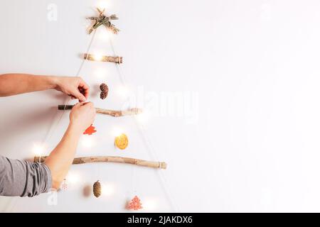 Le mani dell'uomo decorano l'albero di Natale fatto dai bastoni asciutti, arancione asciutto, coni su sfondo bianco. Zero rifiuti Natale, Capodanno, eco-friendly concep Foto Stock