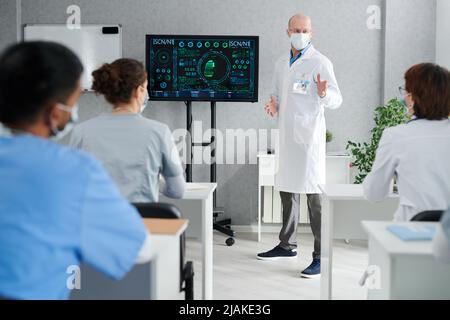 Professore di cappotto bianco e maschera che dà lezioni sull'innovazione medica per gli stagisti presso le scrivanie a classe Foto Stock