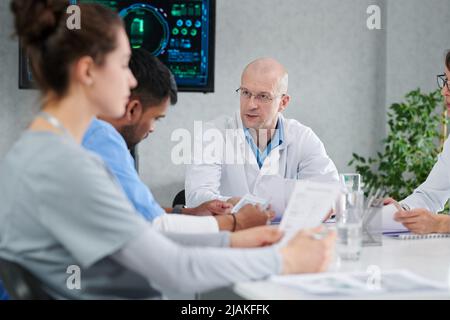 Medico maschile maturo in cappotto bianco seduto a tavola con i suoi colleghi e discutere di lavoro durante l'incontro in ufficio Foto Stock