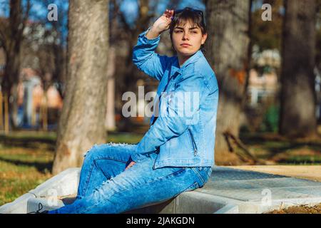 Bella ragazza con corti capelli neri nel suo 20s nel parco che indossa jeans blu chiaro e una giacca di denim blu chiaro Foto Stock