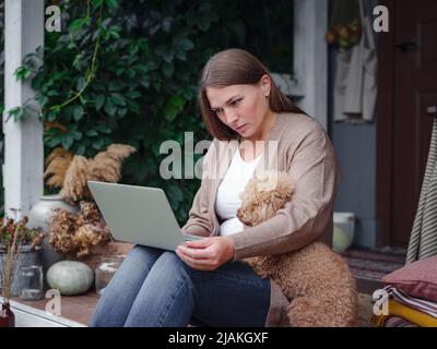 donna caucasica di mezza età in cardigan marrone si siede sulla terrazza di casa con il suo barile beige con il laptop che lavora all'aperto in giardino, ufficio domestico, istruzione, stile di vita moderno e concetto di svago. Foto Stock