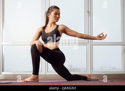 Vista frontale di una donna graziosa e flessibile seduta sul tappeto yoga in posa bassa di yoga in studio. Ragazza snella in leggings neri che posano con il braccio, stirata in aria con il gesto compiuto. Concetto di yoga. Foto Stock