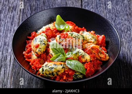 Malfatti, gnocchi di ricotta di spinaci italiani in salsa di pomodoro con erbe e parmigiano grattugiato in ciotola nera su tavola di legno scuro Foto Stock