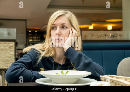 Giovane donna bionda solitaria e triste in un bar con un piatto di cibo. Messa a fuoco selettiva. Foto Stock
