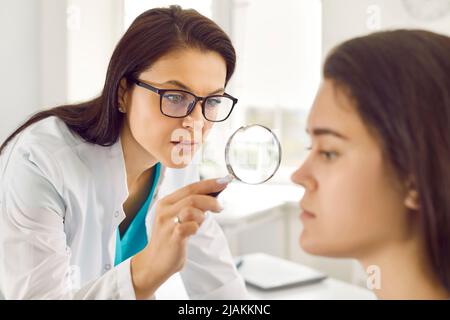 Dermatologo utilizza lente d'ingrandimento per esaminare la pelle problema sul viso della giovane donna Foto Stock
