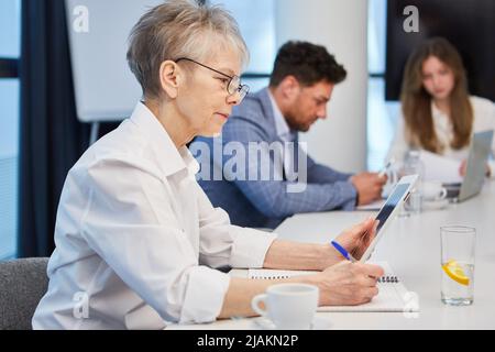 Una donna d'affari anziana siede in una riunione e si concentra sul suo tablet PC Foto Stock