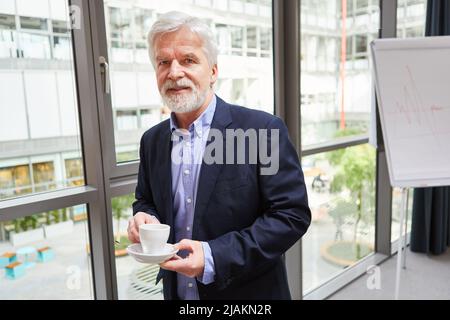 L'uomo d'affari anziano è in piedi dalla finestra in ufficio durante una pausa e bere una tazza di caffè Foto Stock