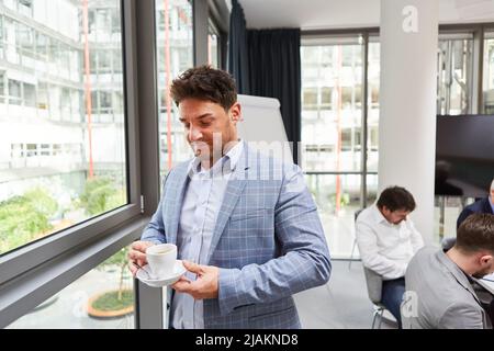 L'uomo d'affari si rilassa bevendo una tazza di caffè durante una pausa accanto alla finestra in ufficio Foto Stock
