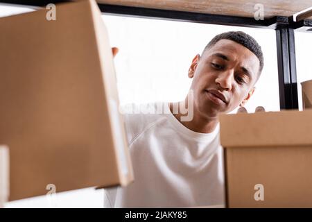 Venditore afroamericano che guarda i pacchetti blurred sulla cremagliera nel deposito in linea di fotoricettore Foto Stock