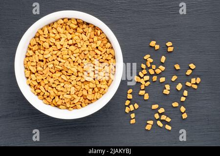 Fenugreek (Trigonella foenum-graecum) in ciotola di ceramica e mazzo su sfondo di legno nero. Macro. Disposizione piatta. Concetto di cibo vegetariano Foto Stock