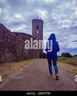 Una signora con la schiena girata indossando un cappotto blu con cappuccio che cammina verso una chiesa sotto il cielo nuvoloso a Monells Foto Stock