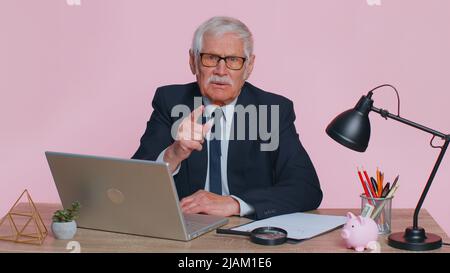 Uomo d'affari senior in scuote il dito e dire di no, fare attenzione, scolding e dare consigli per evitare il pericolo errore, segno di disapprovazione in ufficio. Uomo anziano isolato da solo su sfondo rosa studio parete Foto Stock