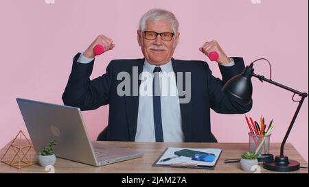 Uomo d'affari anziano che si sta allenando, pompando i muscoli del braccio che sollevano manubri rosa, rilassandosi dopo il lavoro, assistenza sanitaria in ufficio. Motivazione sportiva per l'uomo anziano isolato al coperto su sfondo rosa studio Foto Stock