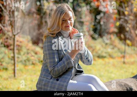 La bionda felice con lunghi capelli ondulati in giacca blu grigia tweed tiene in mano una tazza di caffè riciclabile mentre si siede nel parco autunnale. Giorno di sole in Foto Stock