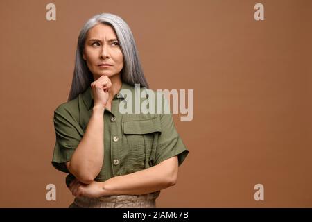 Donna matura intelligente e pensierosa che tiene il mento e ponderante idea, che prende la decisione difficile, che guarda incerta dubbia. Studio interno girato isolato su sfondo beige Foto Stock