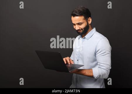 Indiano barbuto ragazzo freelancer utilizzando computer portatile isolato su sfondo nero, uomo multirazziale 30s con capelli scuri in casual shirt dattilografia Foto Stock