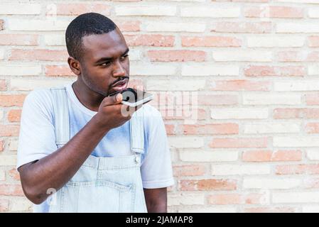 Ragazzo afroamericano serio e giovane in abiti casual. Sta registrando un messaggio vocale sullo smartphone e guardando lontano mentre si trova vicino al muro di mattoni sulla strada della città Foto Stock