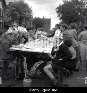 1953, storico, festa di strada che celebra l'incoronazione della regina Elisabetta II al trono, un gruppo di persone sedute a tavola in una strada, con ragazzi alla fine del tavolo, seduti con i cappelli, alcuni con la lettera e su, Inghilterra, Regno Unito. Foto Stock