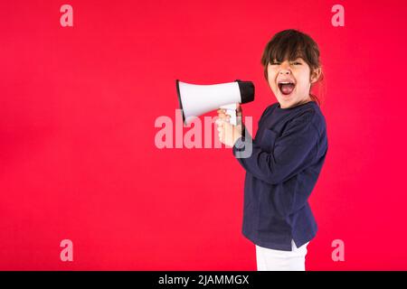 Ragazza dai capelli neri in un maglione blu e pantaloni bianchi, gridando attraverso un megafono bianco, su sfondo rosso. Foto Stock