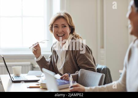 Felice insegnante donna tenendo occhiali guardando un collega in stafroom Foto Stock