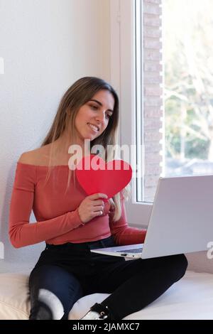 Ritratto di una giovane ragazza che fa una videocall con il suo ragazzo tenendo il cuore rosso in una data di coppia. San Valentino in rapporti a lunga distanza Foto Stock