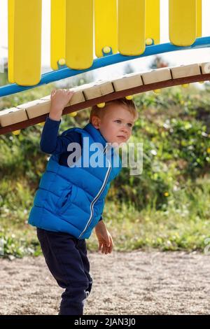 Un ragazzo piccolo carino gioca sotto un bel ponte curvo su un colorato parco giochi. Un bambino agile trascorre attivamente del tempo all'aria aperta. Puntino verticale Foto Stock