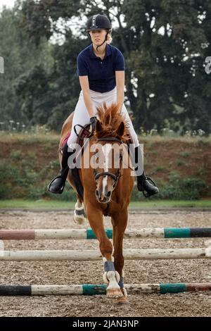 Cavaliere equestre - giovane ragazza che salta con cavallo marrone ciliegia. Un cavallo e un cavaliere in moto di salto, in aria, competizione equestre sotto la pioggia. Foto Stock